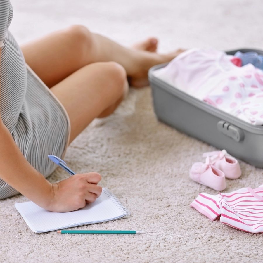 Future maman écrivant sa liste de naissance à côté d'une valise de maternité